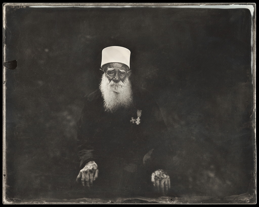 monochrome photograph of an elderly Druze man, seated on the floor with his hands on his knees